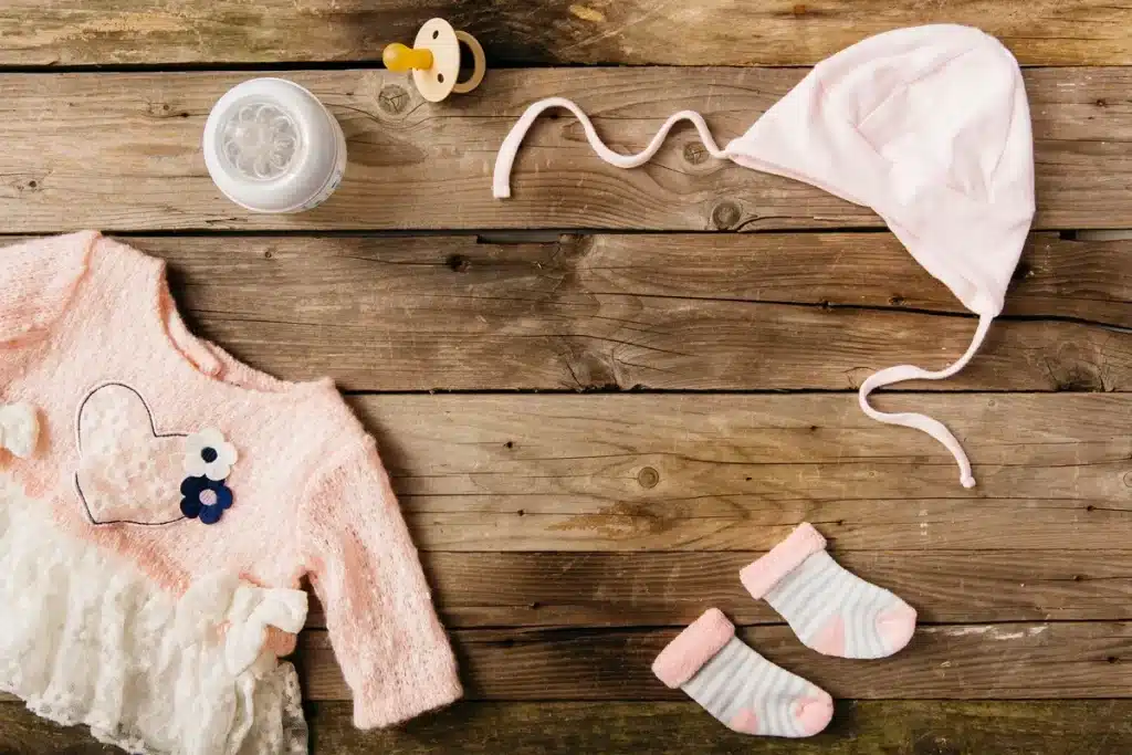 Conjunto rosa para bebê menina, com um suéter decorado, meias listradas, chupeta e um chapéu, tudo sobre uma mesa de madeira.