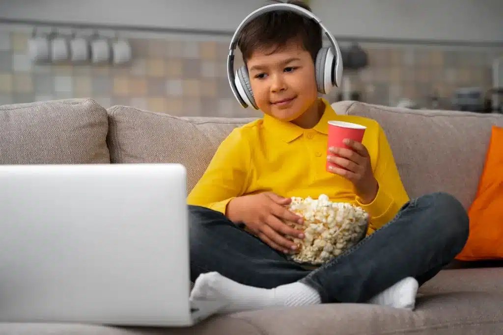 Criança assistindo a uma série infantil no laptop com fones de ouvido, segurando pipoca e refrigerante.