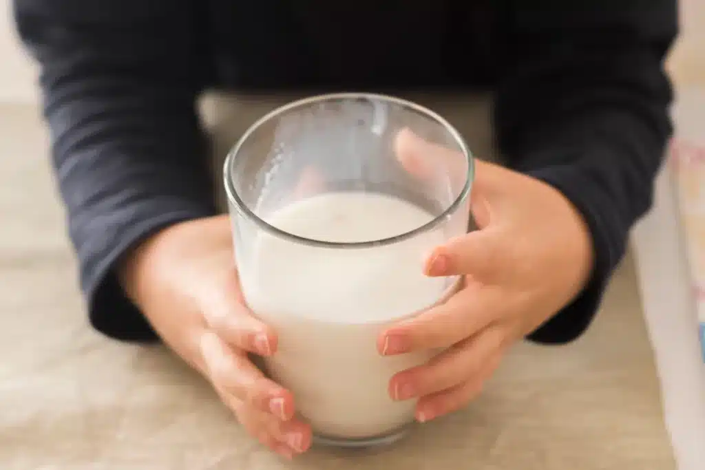 Criança segurando um copo de leite, com as mãos delicadamente envoltas no vidro. A imagem sugere a ingestão de leite, que pode causar sintomas de intolerância à lactose, como dor abdominal e desconforto digestivo.
