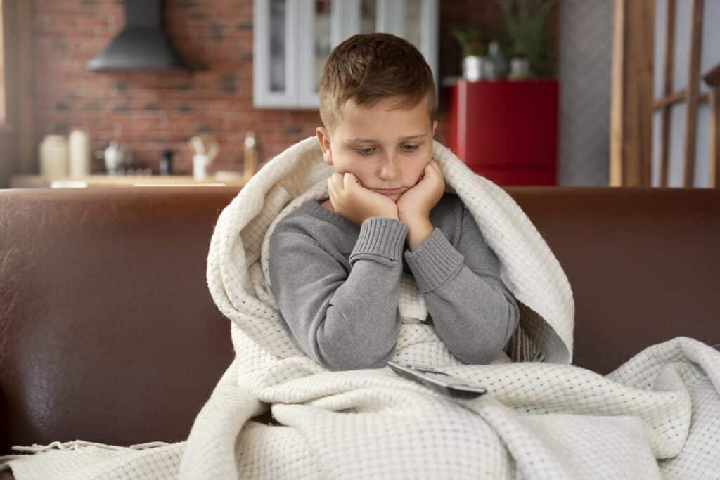 Criança assistindo TV no sofá com expressão triste, coberta por um cobertor branco e segurando o controle remoto.