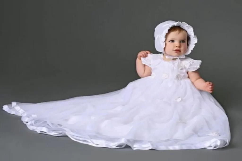 Fotografia de estúdio de um bebê menina (com aproximadamente 6 a 12 meses) sentado em um fundo cinza-sólido. A bebê veste um vestido de festa ou batizado branco, com mangas bufantes, detalhes de flores em relevo e uma cauda longa de tule que se espalha pelo chão. Ela também usa uma touca (capuz) branca combinando com babados na borda. A roupa é formal e delicada, ideal para cerimônias infantis.