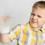 Criança segurando um copo de leite com uma expressão de desgosto, possivelmente indicando uma reação a sintomas de intolerância à lactose, como cólicas ou desconforto abdominal.