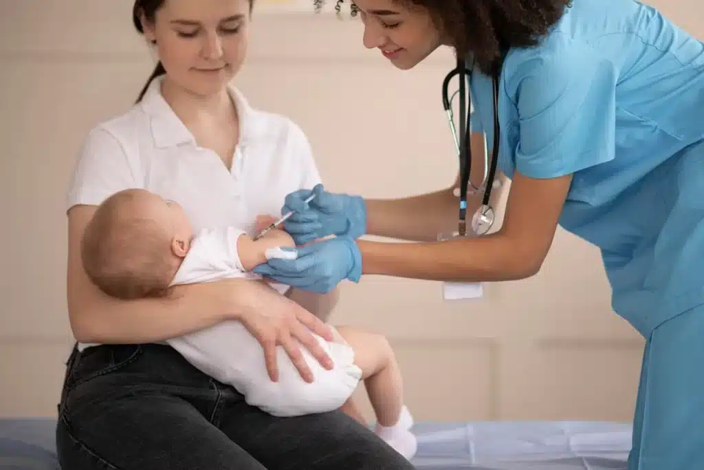 Enfermeira aplicando vacina no braço de um bebê no colo da mãe durante atendimento em consultório.