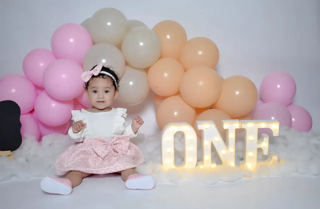 Bebê comemorando o primeiro aniversário com decoração delicada em tons de rosa, creme e pêssego, com balões coloridos e letras iluminadas formando a palavra 'ONE'.