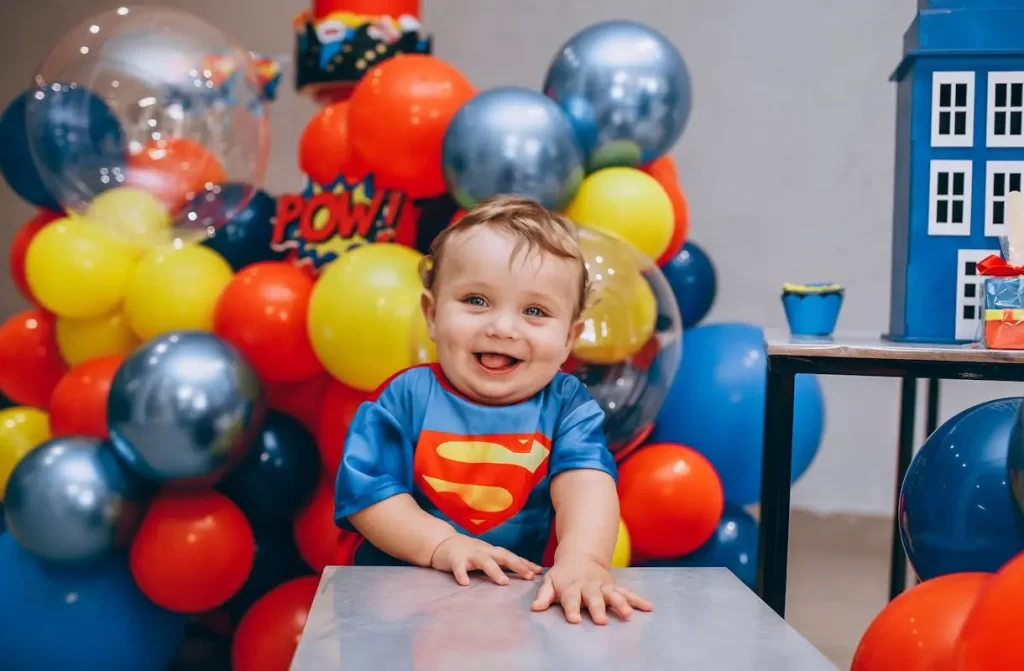 Bebê sorridente vestido de Super-Homem em festa de um ano com decoração temática de super-heróis. Ao fundo, balões coloridos nas cores vermelho, amarelo, azul e prata, além de elementos gráficos como 'POW!' em estilo de quadrinhos.