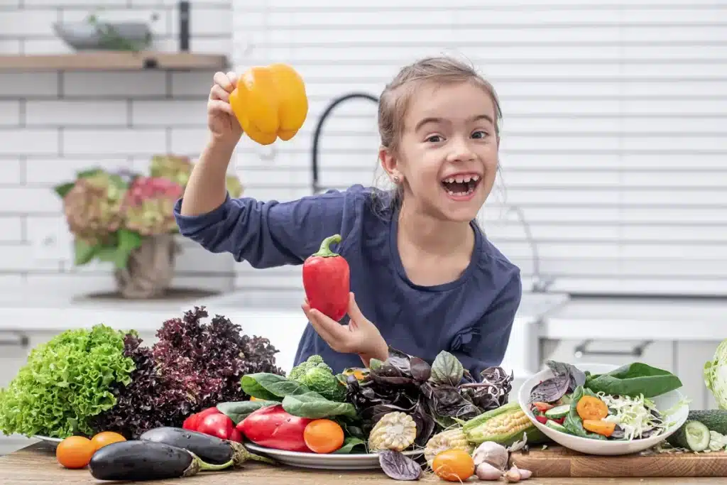 Menina sorrindo com pimentões vermelho e amarelo, rodeada por vegetais frescos como alface, pepino e berinjela