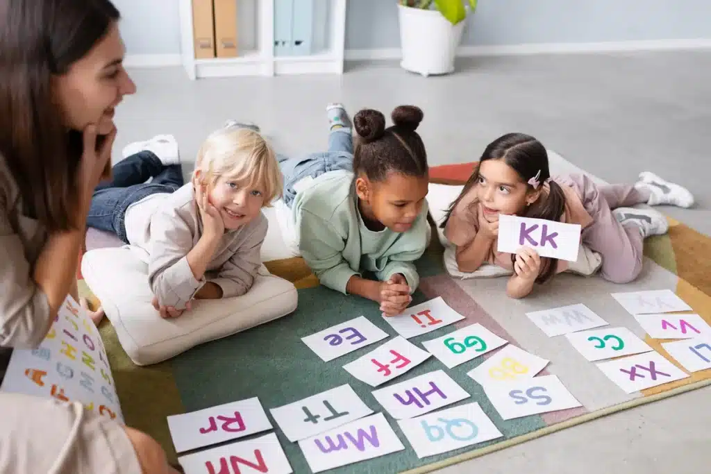 Crianças aprendendo o alfabeto com professora em sala de aula divertida e interativa. Atividade educativa com letras e cartões coloridos no chão.