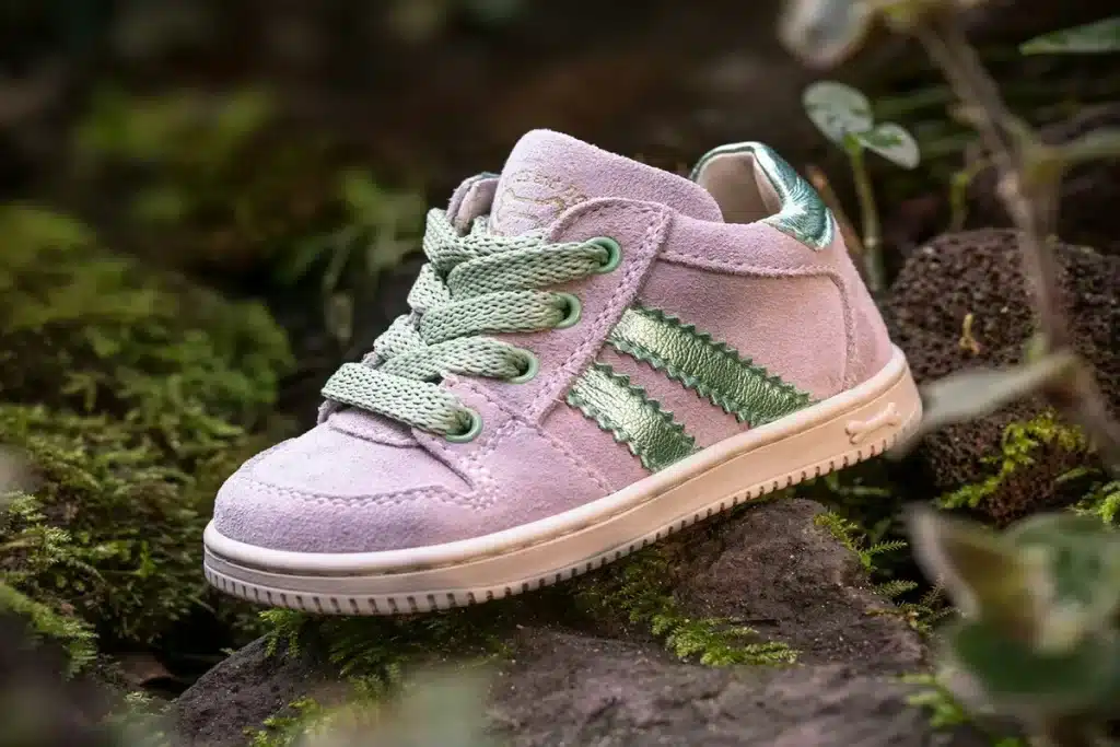 Tênis infantil rosa claro com detalhes verde metálico, em destaque sobre fundo natural com musgos e pedras.