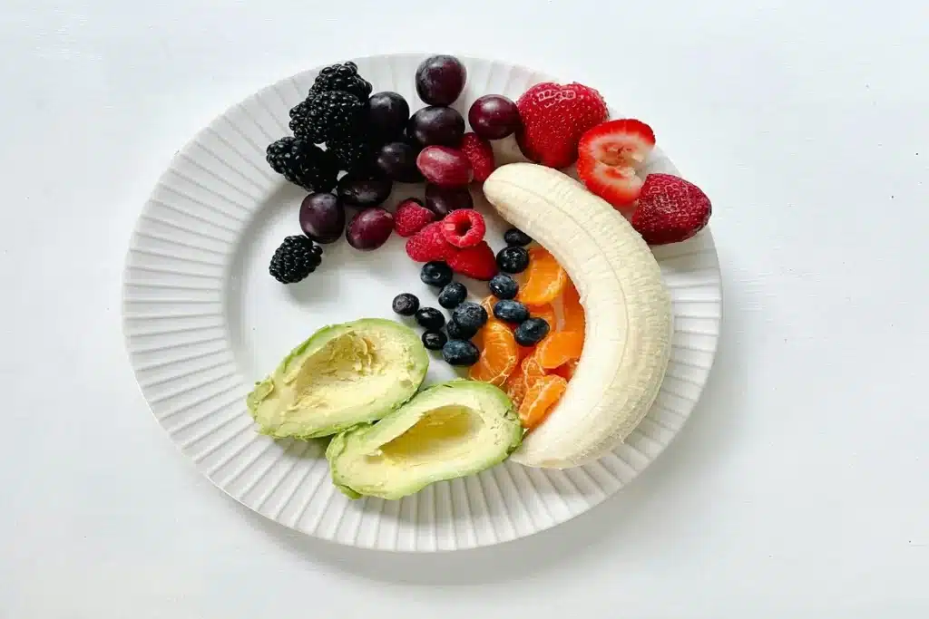 Prato branco com banana, morango, uva, framboesa, amora, mirtilo, tangerina e abacate, representando uma refeição colorida e saudável ideal para dieta vegetariana