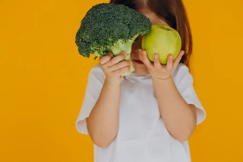 Menina segurando brócolis e maçã verde na frente de fundo amarelo, promovendo alimentos saudáveis