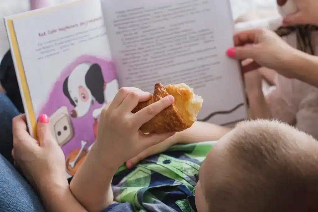 Criança deitada segurando um livro infantil ilustrado com cachorro e comendo um croissant