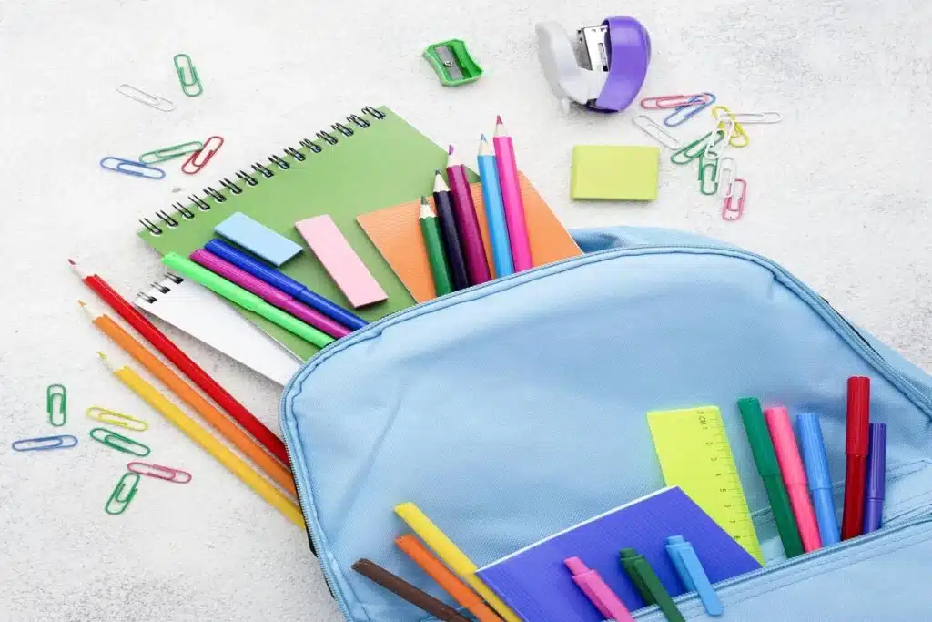 Mochila azul aberta com diversos itens da lista de material escolar, incluindo cadernos, lápis de cor, canetas coloridas, borrachas, régua, grampeador e clipes, organizados sobre uma superfície clara.