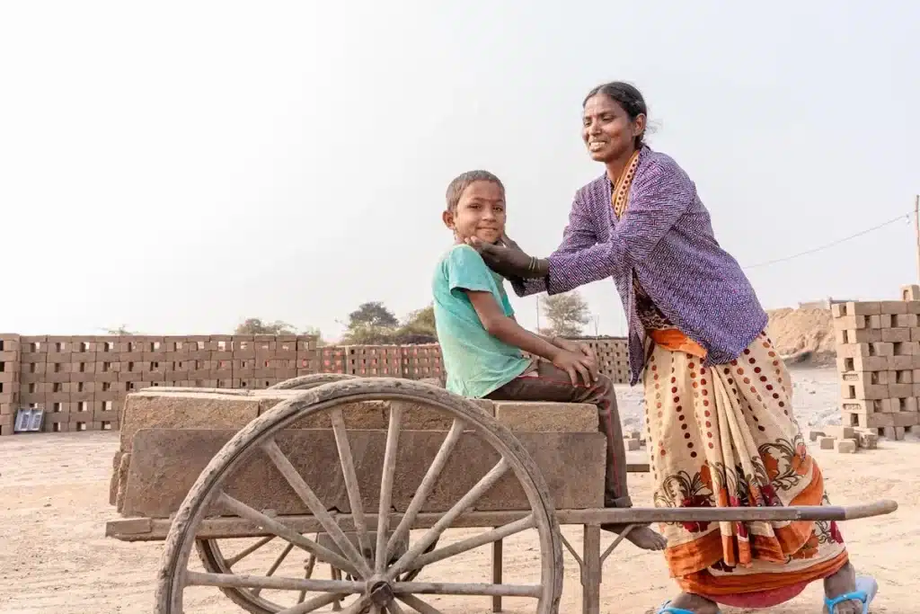Mulher sorrindo em trajes indianos, tocando o rosto de um menino que está sentado em uma carroça carregada de tijolos em um canteiro de obras ou olaria.