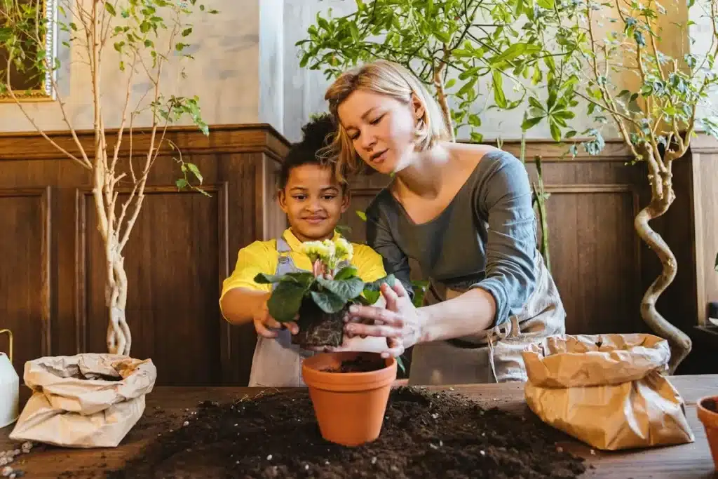 Mulher e criança pequena de cabelos cacheados replantando uma flor em um vaso de terracota, com as mãos na terra, em uma atividade de jardinagem.