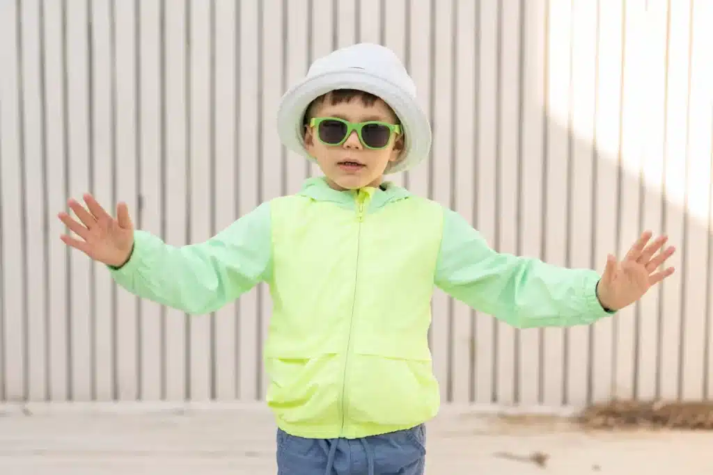 Criança usando jaqueta neon verde-limão, óculos de sol verdes e chapéu branco, posando com os braços abertos em frente a parede listrada