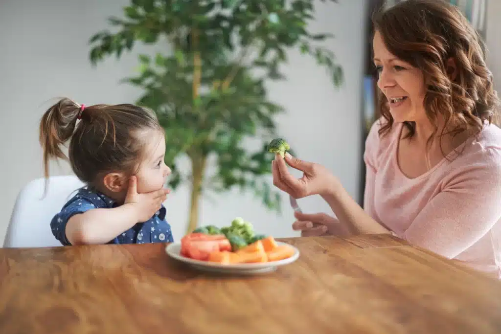 Mãe oferecendo brócolis para filha pequena com seletividade alimentar durante refeição saudável em casa.