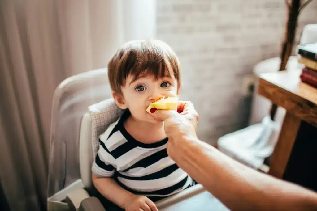 Criança pequena sendo alimentada com pedaço de maçã por uma mão, com expressão curiosa e atenta, usando uma camiseta listrada.