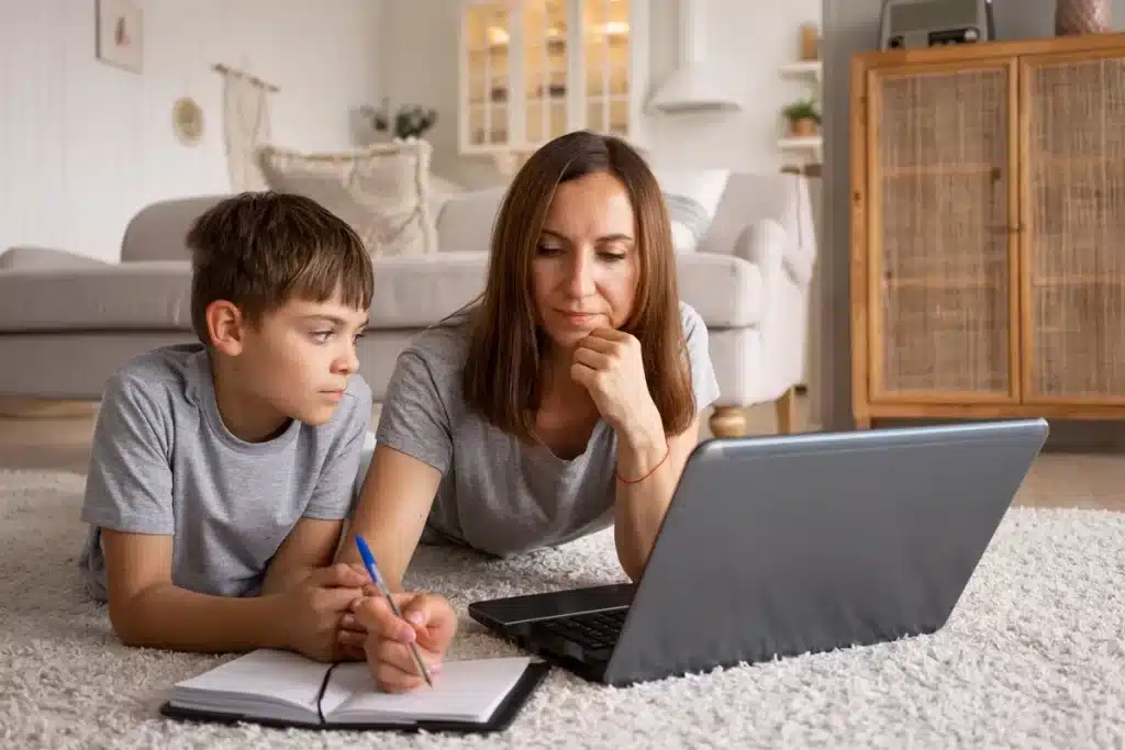 Imagem de uma mãe e seu filho estudando no chão, com o filho escrevendo em um caderno enquanto a mãe observa atentamente a tela do laptop.