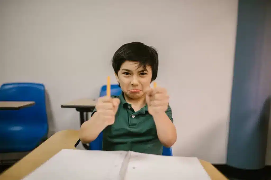 Menino na carteira escolar fazendo careta de raiva ou frustração, segurando lápis nas mãos. Pode indicar desafios relacionados ao "Transtorno Explosivo Intermitente" ou dificuldades de aprendizado.