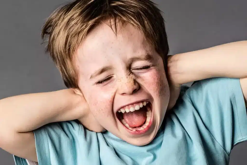 Criança com sardas e cabelos bagunçados gritando com as mãos nos ouvidos, expressando angústia ou explosão de raiva, ilustrando o "Transtorno Explosivo Intermitente" (TEI).