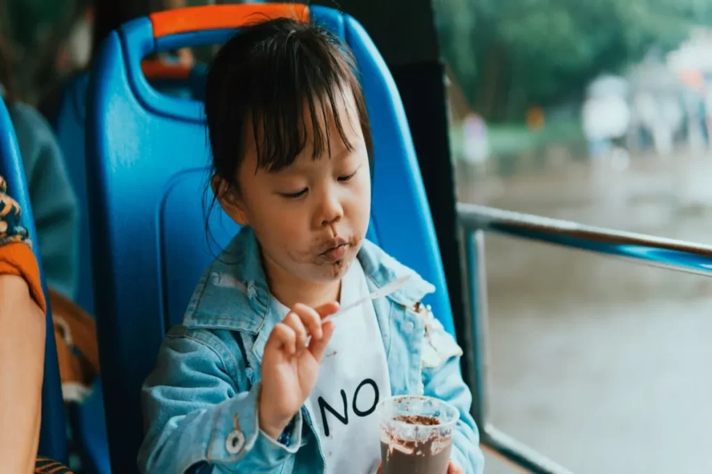 Uma criança pequena, sentada num assento azul de autocarro, comendo algo de um copo com uma colher, com o rosto sujo de chocolate. A imagem representa a infância, a pureza e as características desta fase da vida.
