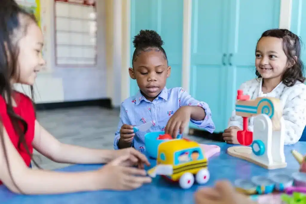 Três meninas de diferentes etnias brincando juntas com brinquedos como um kit de cozinha infantil e um carrinho escolar amarelo. A imagem sugere "brinquedos de meninas" que estimulam a imaginação e o faz de conta.