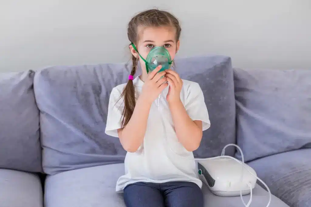 Menina usando nebulizador com máscara para ajudar a descongestionar o nariz, sentada em um sofá.