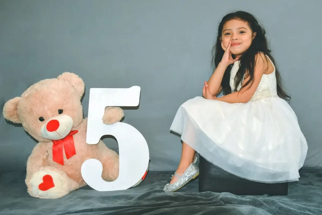 Menina sorridente de cinco anos sentada ao lado de um urso de pelúcia e um número cinco grande, representando uma das adoráveis fases da infância.