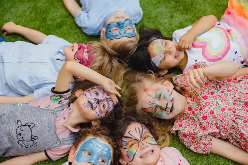 Grupo de crianças com pintura facial de animais e super-heróis deitado na grama durante festa infantil ao ar livre