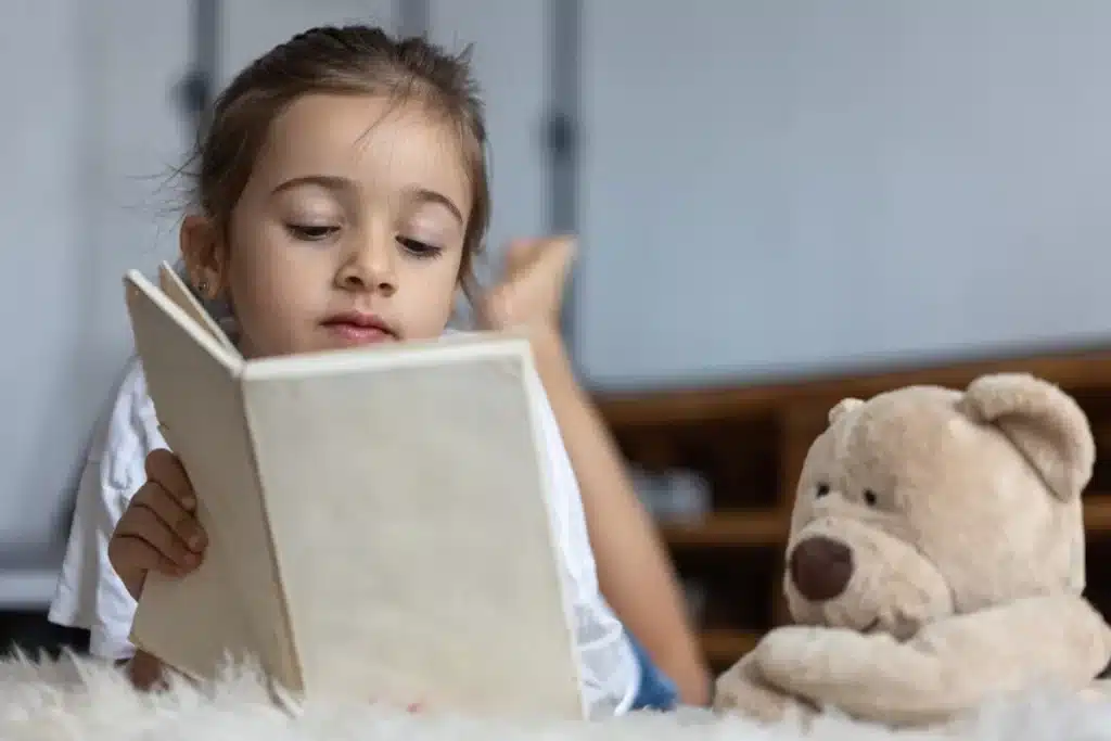 Uma menina pequena, deitada de bruços em um tapete felpudo e branco, lê um livro de capa bege com concentração, enquanto um grande urso de pelúcia marrom está deitado ao seu lado, ilustrando a leitura de histórias curtas.