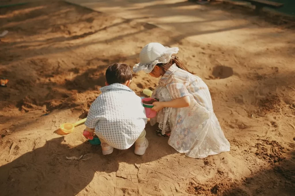 Duas crianças com baldinho e pá brincando em grande caixa de areia.