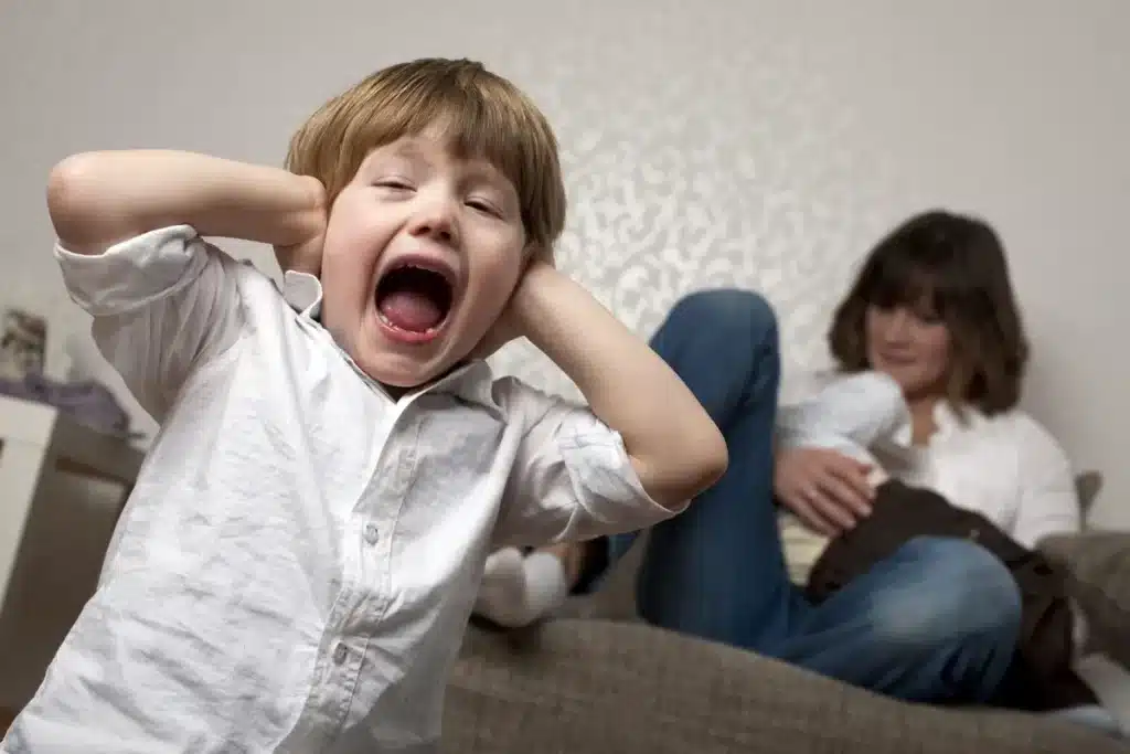 Criança agitada e com as mãos nos ouvidos, menino com comportamento impulsivo, mãe no sofá com um bebê.
