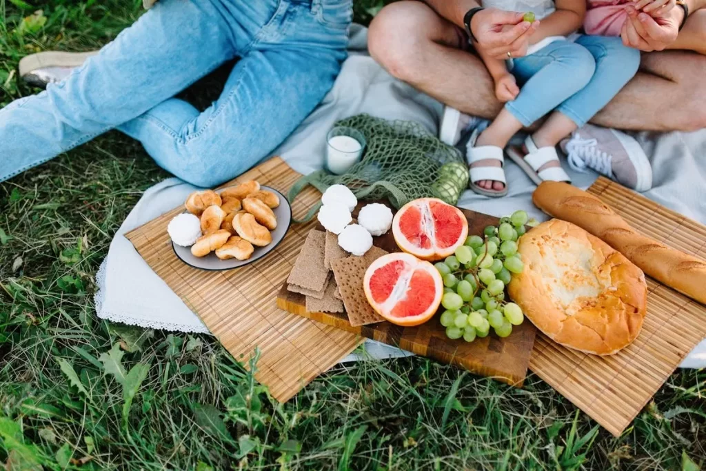 Cesta de piquenique com frutas frescas, pães e doces, disposta sobre um lençol na grama, com pernas de adultos e crianças ao redor, desfrutando um lanche da tarde em família.