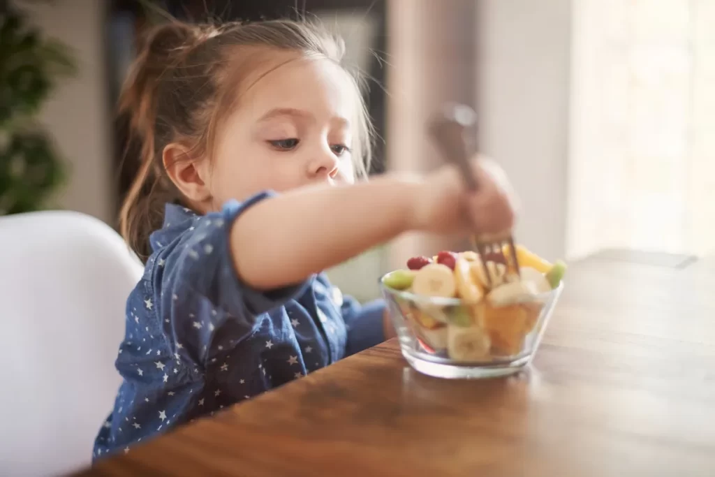 Menina focada, desfrutando de um lanche da tarde saudável com um garfo e tigela de frutas coloridas.