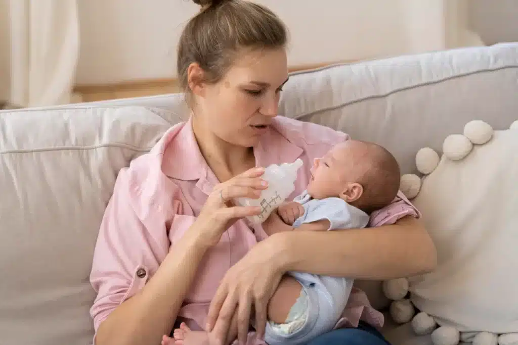 Mãe alimentando recém-nascido com mamadeira, sentada em um sofá com almofadas.