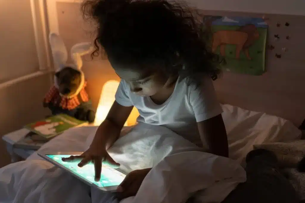 Menina usando tablet no escuro do quarto antes de dormir, tela iluminando o rosto, uso de tablet infantil, entretenimento noturno.