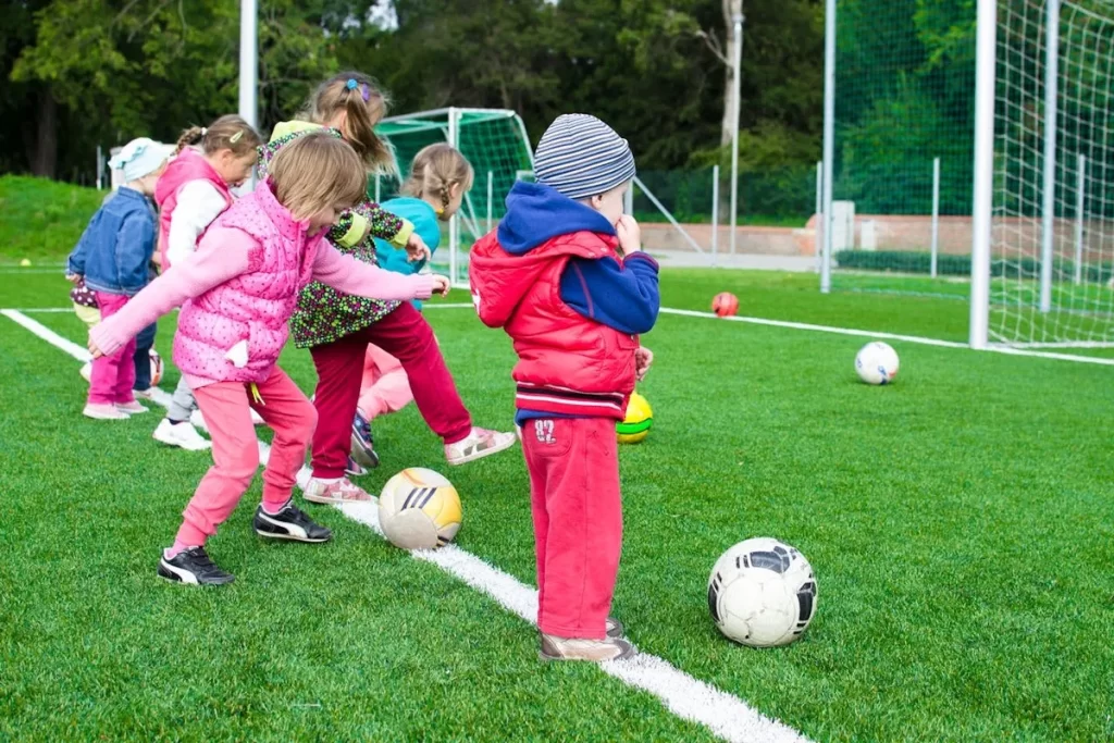 Grupo de crianças pequenas brincando de futebol em um campo de grama verde, algumas chutando a bola e uma criança em primeiro plano observando, representando a diversão e a importância da atividade física para todas as crianças, incluindo aquelas com "mobilidade reduzida".