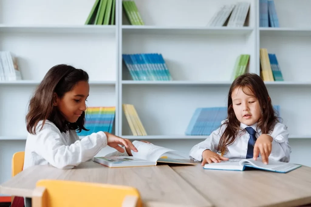 Duas meninas em uniforme escolar lendo livros em mesa de madeira, com estantes cheias de livros ao fundo, em um cantinho de leitura escolar no Brasil.