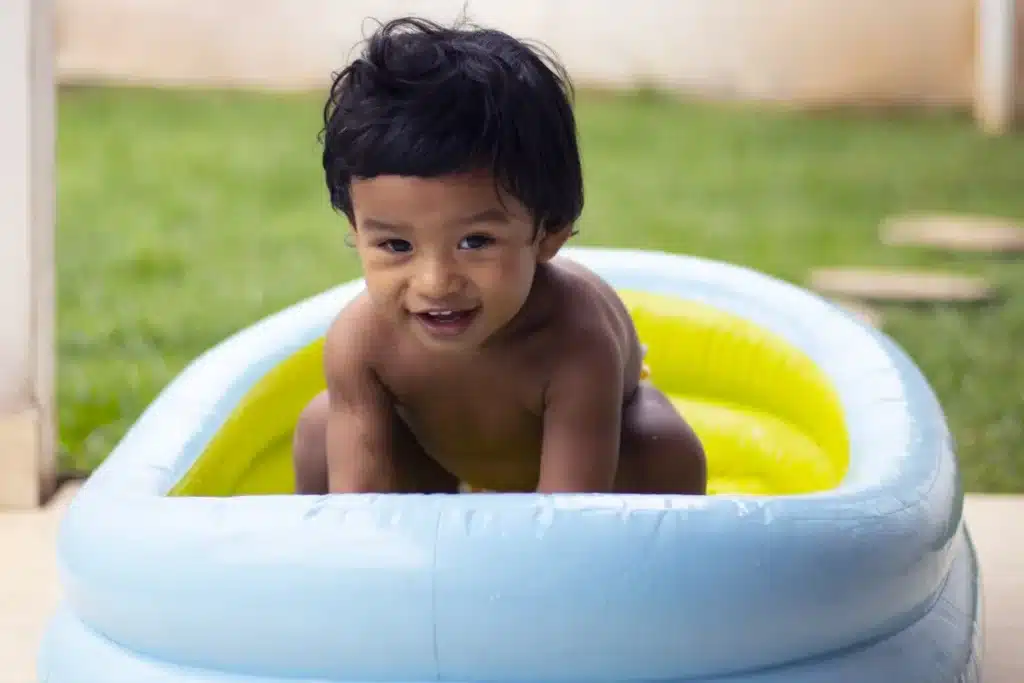 Bebê sorridente brincando em uma piscina inflável azul e amarela no quintal, aproveitando o dia de verão.
