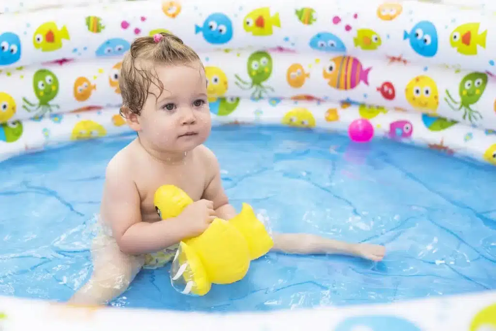Bebê brincando feliz em uma piscina inflável colorida com água e brinquedos, ideal para diversão no verão.