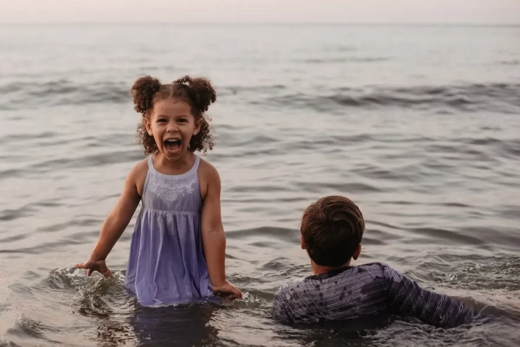 Uma menina pequena com cabelo cacheado, vestindo um vestido lilás, sorrindo e brincando na água do mar ao lado de um menino, ideal para "praia para ir com criança".