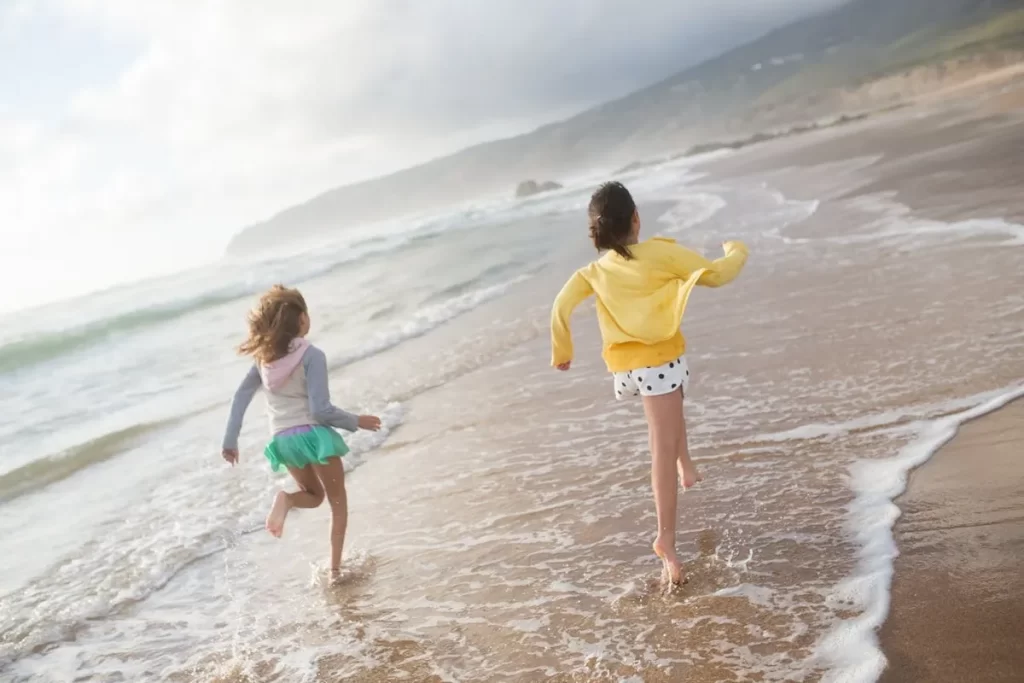 Duas crianças felizes correndo na beira de uma praia ensolarada, com ondas suaves e montanhas ao fundo, ideal para famílias com o tema "praia para ir com criança".