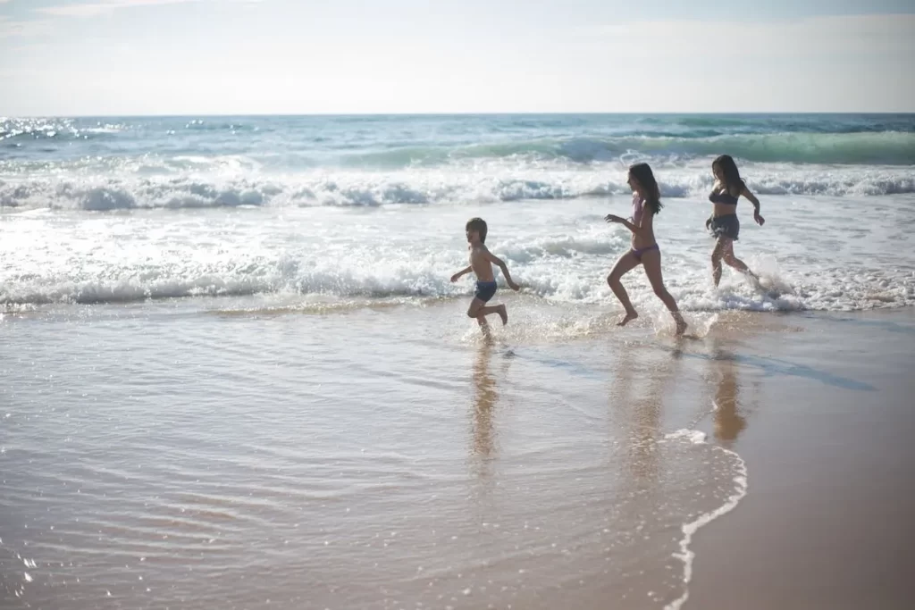 Três crianças de diferentes idades correndo e brincando na água da praia em um dia ensolarado, com o oceano azul e ondas suaves ao fundo, perfeito para o tema "praia para ir com criança".