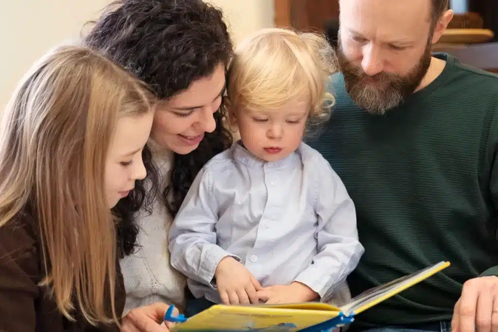 Pai, mãe e dois filhos lendo um livro colorido juntos em casa, promovendo momentos de conexão e aprendizado em família.