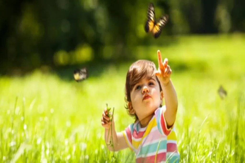 Criança pequena em um campo verde estendendo a mão para pegar uma borboleta, em uma cena que remete a sonhar com criança.