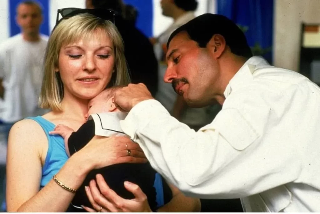 Fotografia de Mary Austin com um bebê no colo e Freddie Mercury em um momento íntimo.