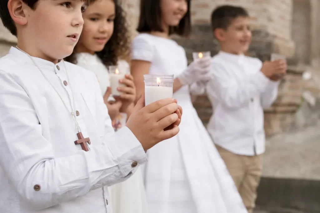 Quatro crianças com roupas brancas, segurando velas, participando da cerimônia de Primeira Comunhão do lado de fora de uma igreja.