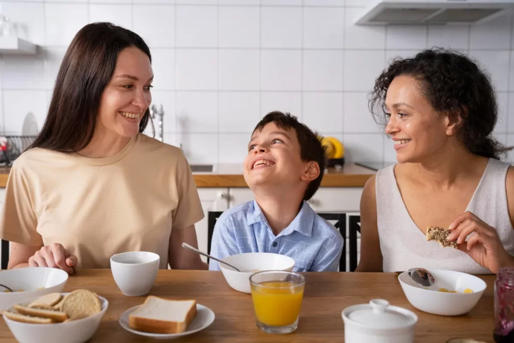 Duas mulheres sorrindo para um menino que está rindo entre elas, durante um almoço em família.