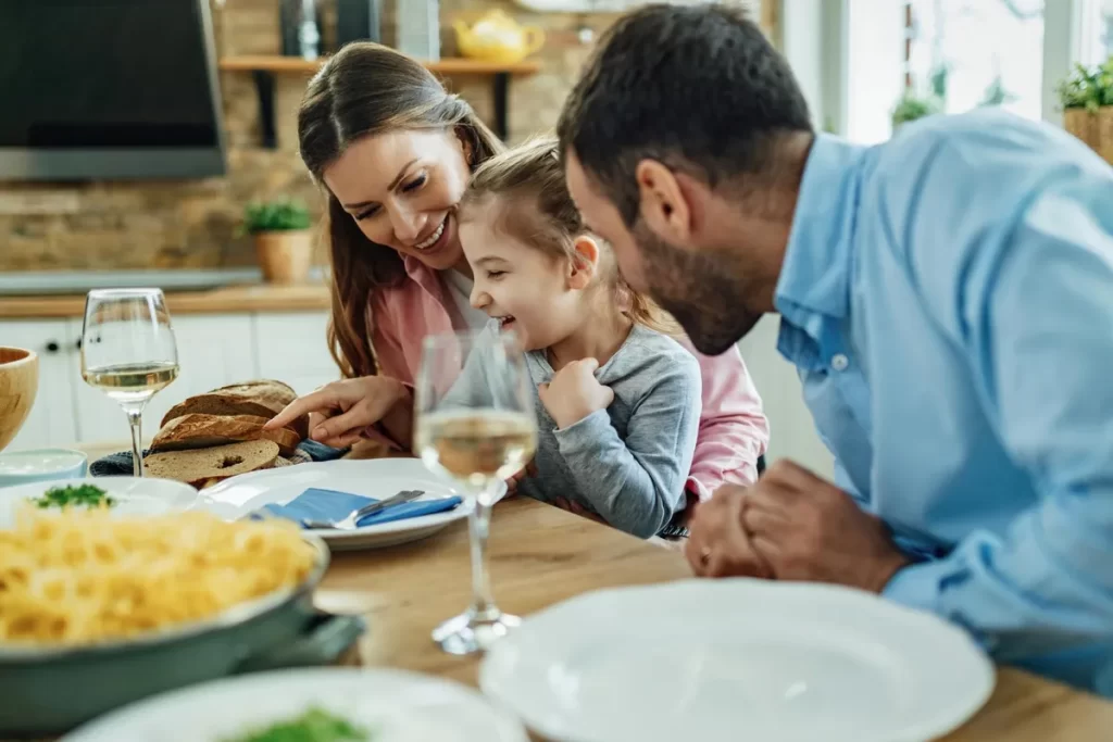 Família feliz em um almoço, com a mãe e o pai sorrindo para a filha. A foto transmite união, alegria e carinho.