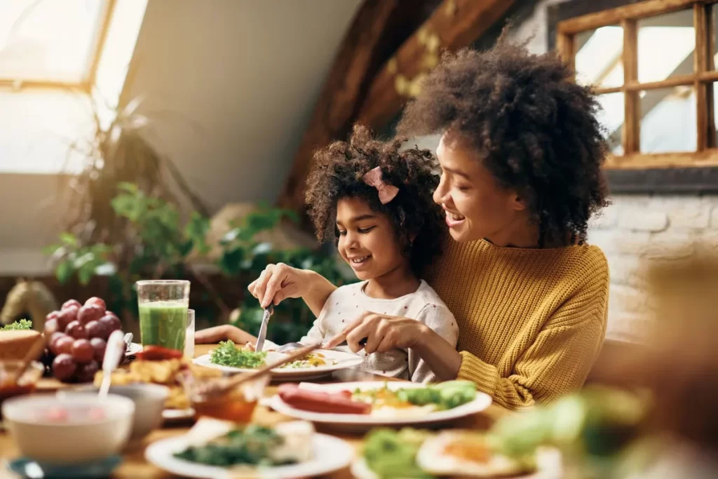 Mãe e filha compartilhando um almoço em família, sorrindo e se divertindo com a refeição. A foto destaca a união, o amor e o carinho familiar.